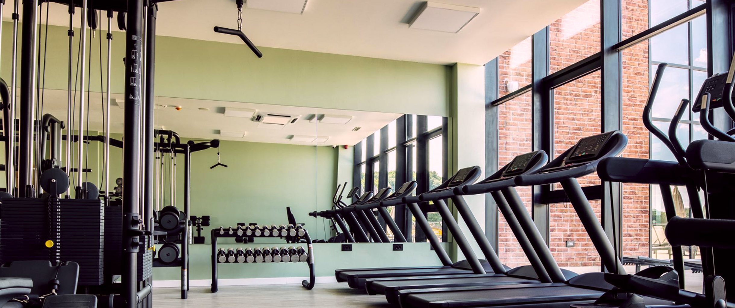 Изображение тренажерного зала в котором с лева блочные тренажеры и гантельные ряды, а с права нескольок беговых дорожек у панорамного окна 